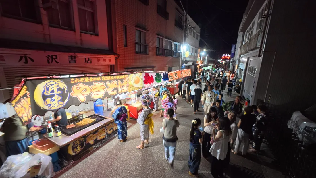 Festival food stalls on opening night: a yakisoba stand with its griddle going, yukata-clad visitors drifting past down the shuttered evening street