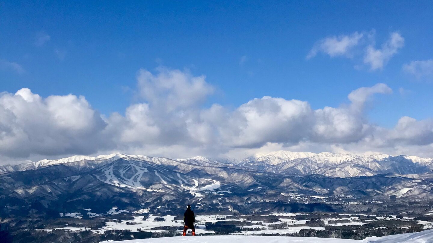 View from the summit of Washigatake ski resort: a lone skier stands on an open snow slope looking out over a vast panorama of Gujo's mountain ranges under a blue sky
