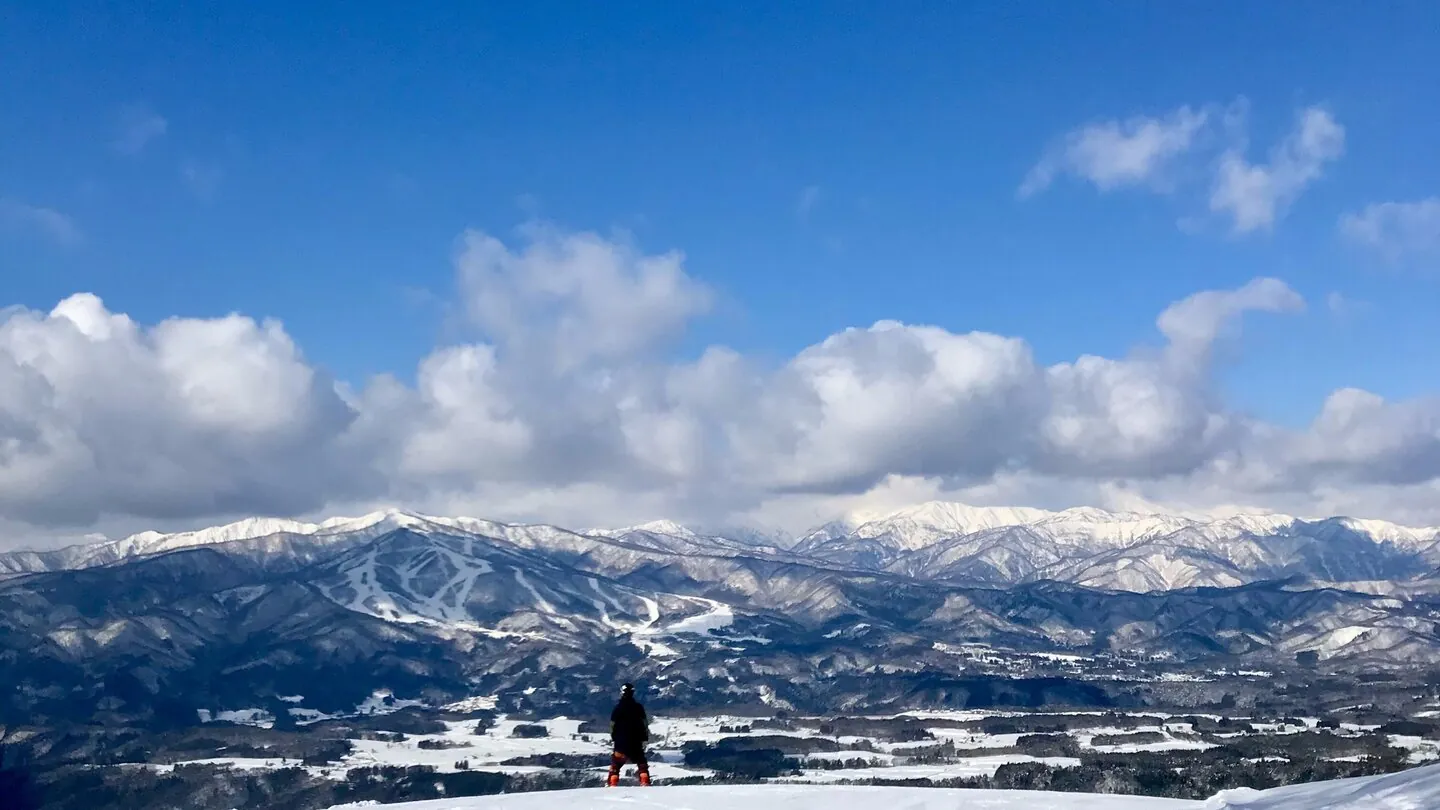 View from the summit of Washigatake ski resort: a lone skier stands on an open snow slope looking out over a vast panorama of Gujo's mountain ranges under a blue sky
